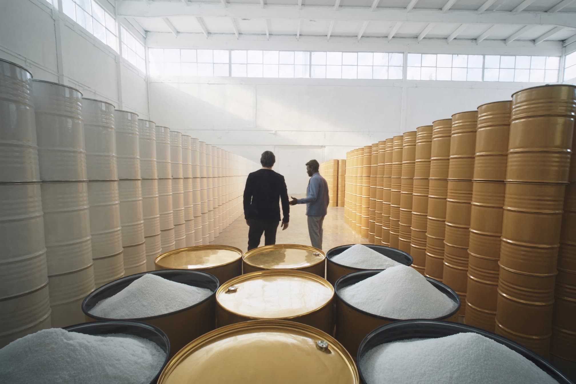 Two people in conversation in an aisle between stacked drums inside a chemical packing hall, with open drums of white crystalline citric acid in the foreground