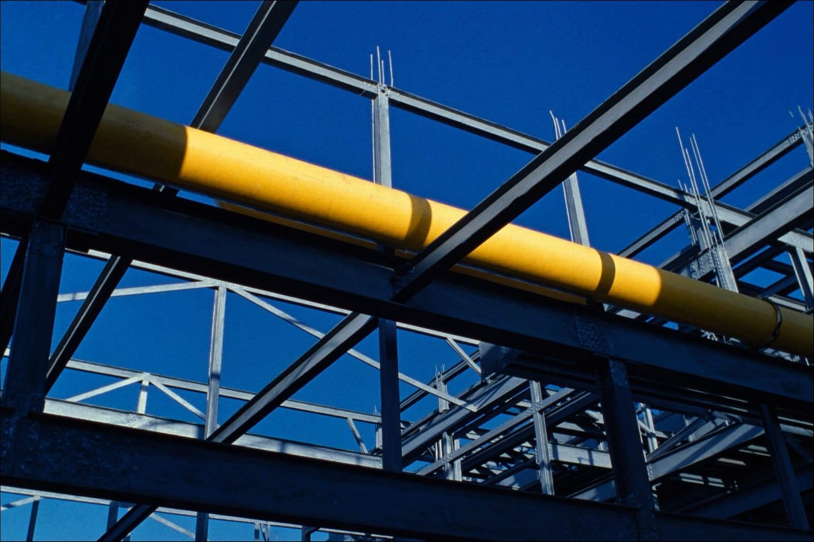 Steel beams crossing overhead against a deep blue sky, a yellow service pipe running through them
