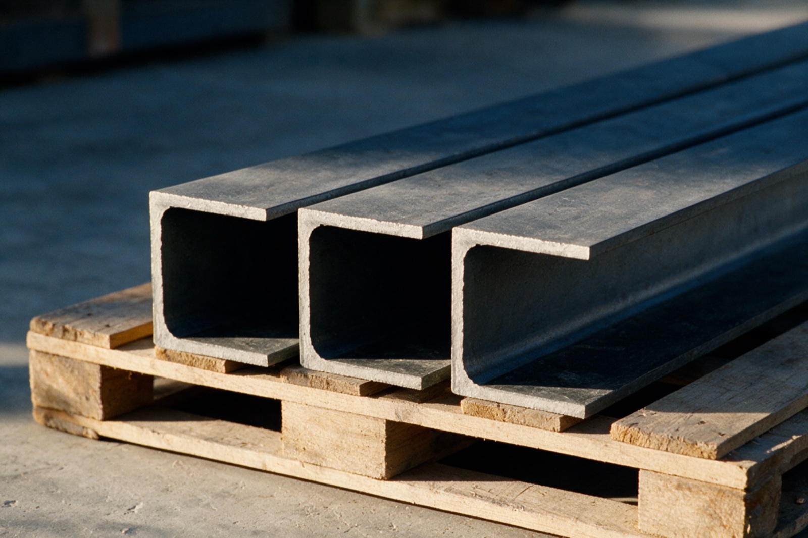Three structural steel channel sections on a timber pallet, their open C profiles facing the camera in low raking light