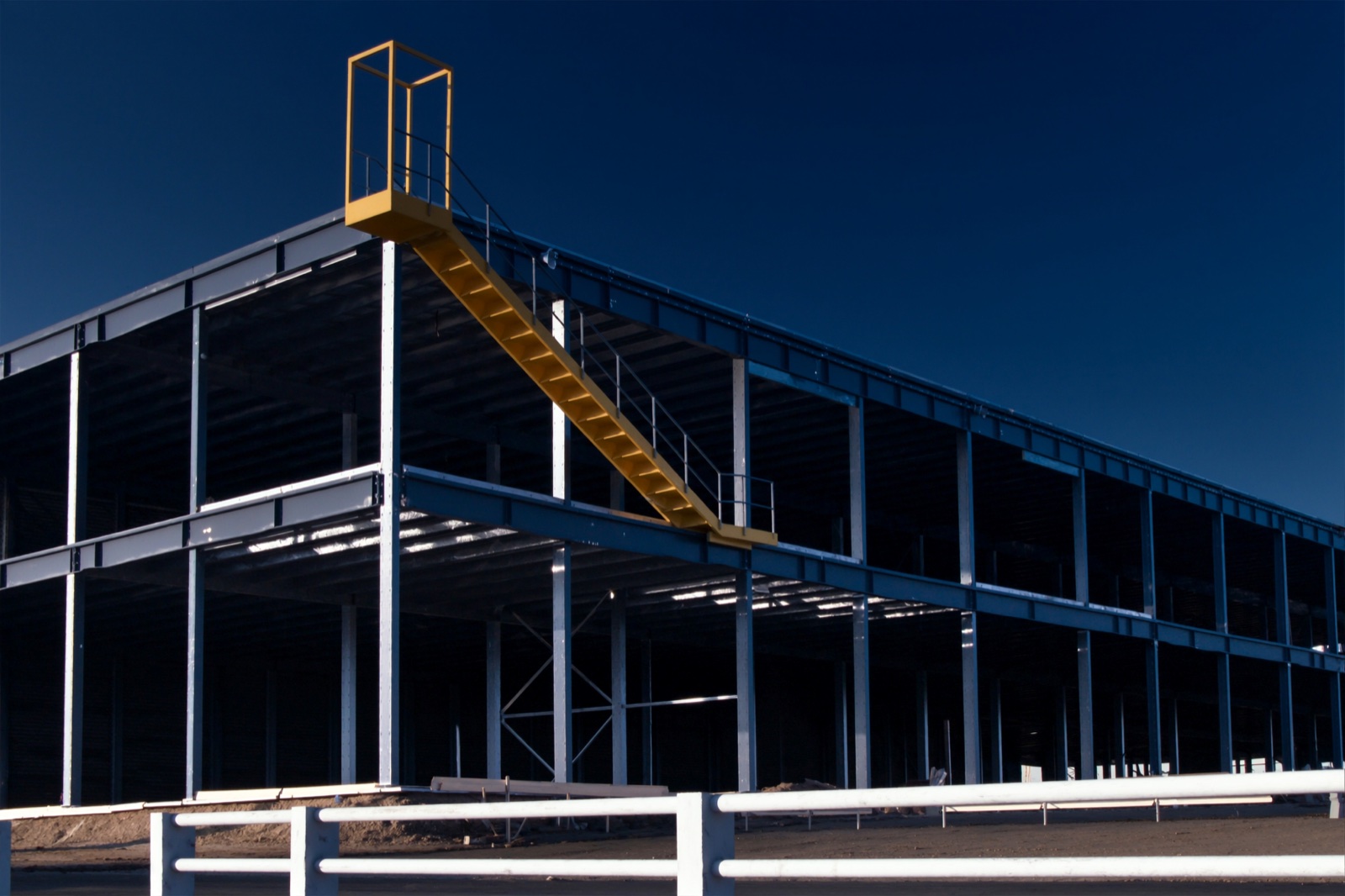 The bare steel frame of a building under construction against a deep blue sky, a yellow access stair running up it