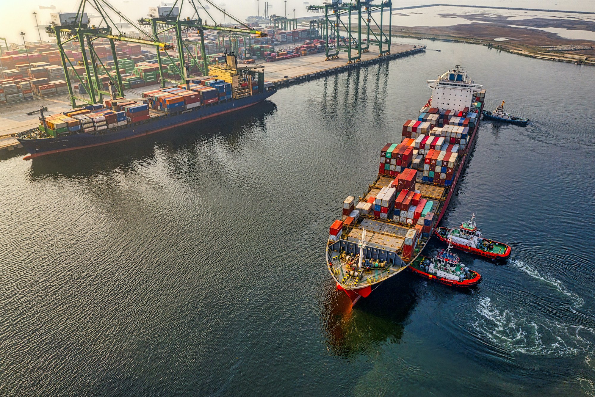 A container vessel under tug escort leaving a port terminal, cranes and stacked containers on the quay