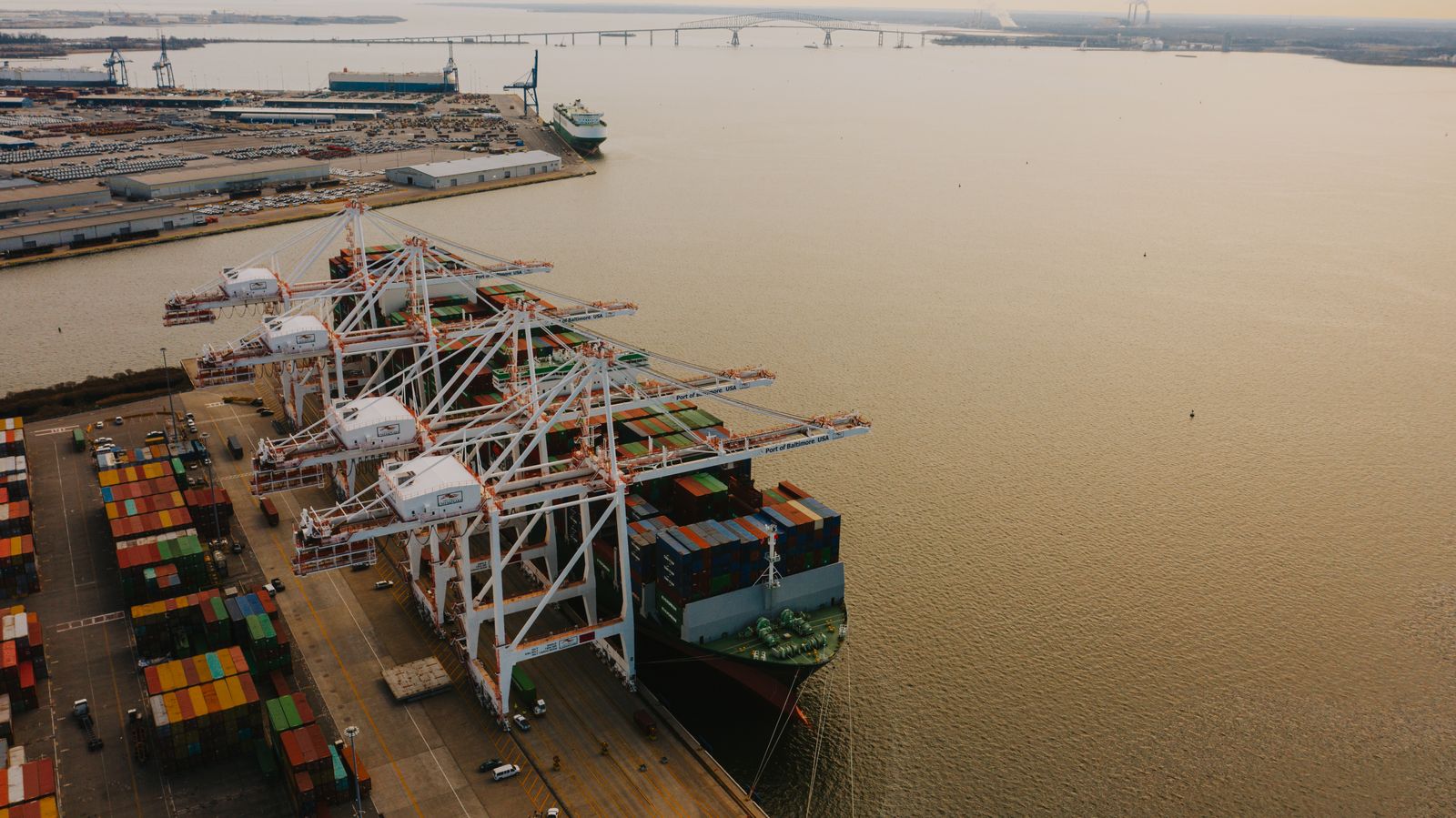 An aerial view of a container terminal at dusk, gantry cranes working a moored vessel beside open water